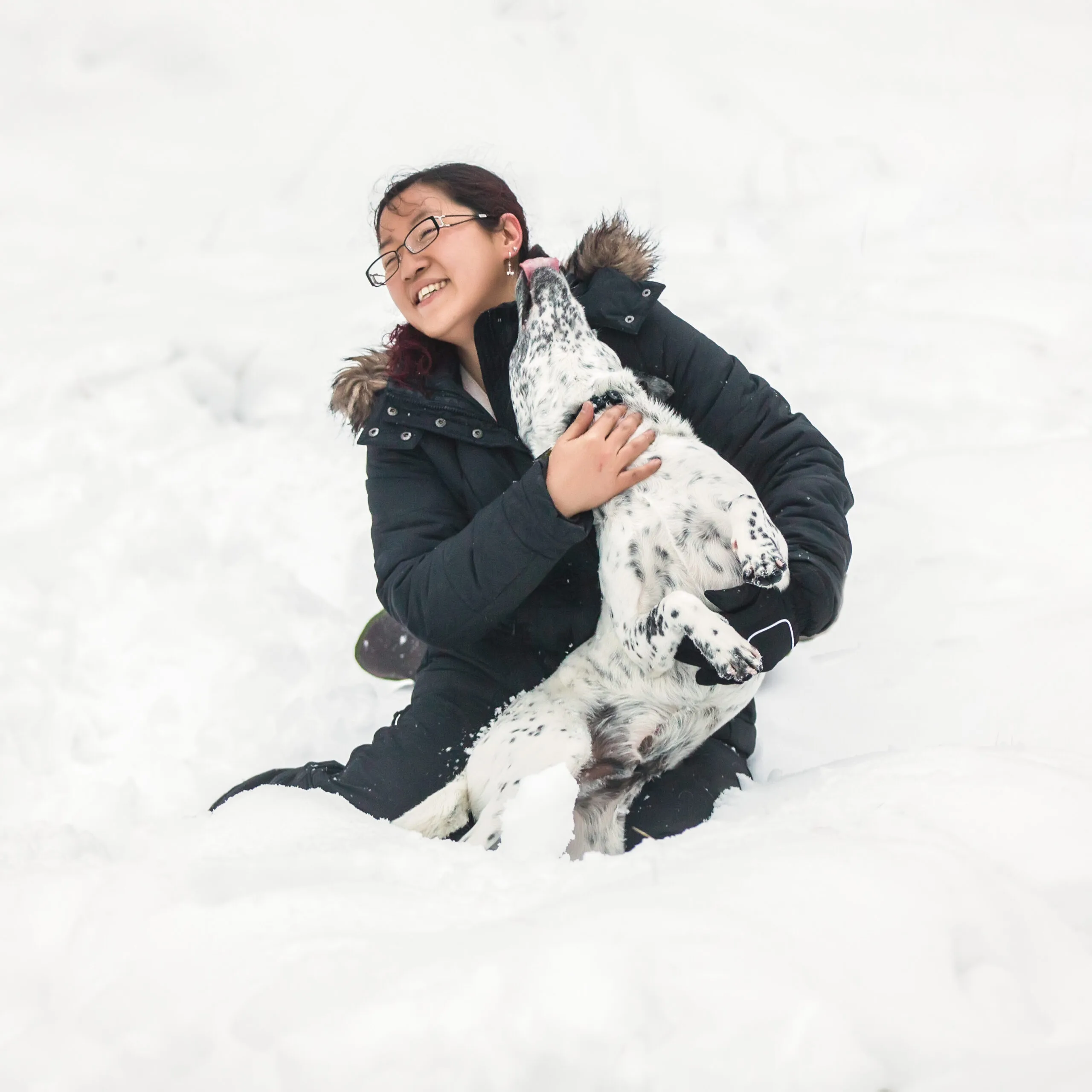 A woman dressed in black snow gear cuddling a  black and white dog while it licks her face