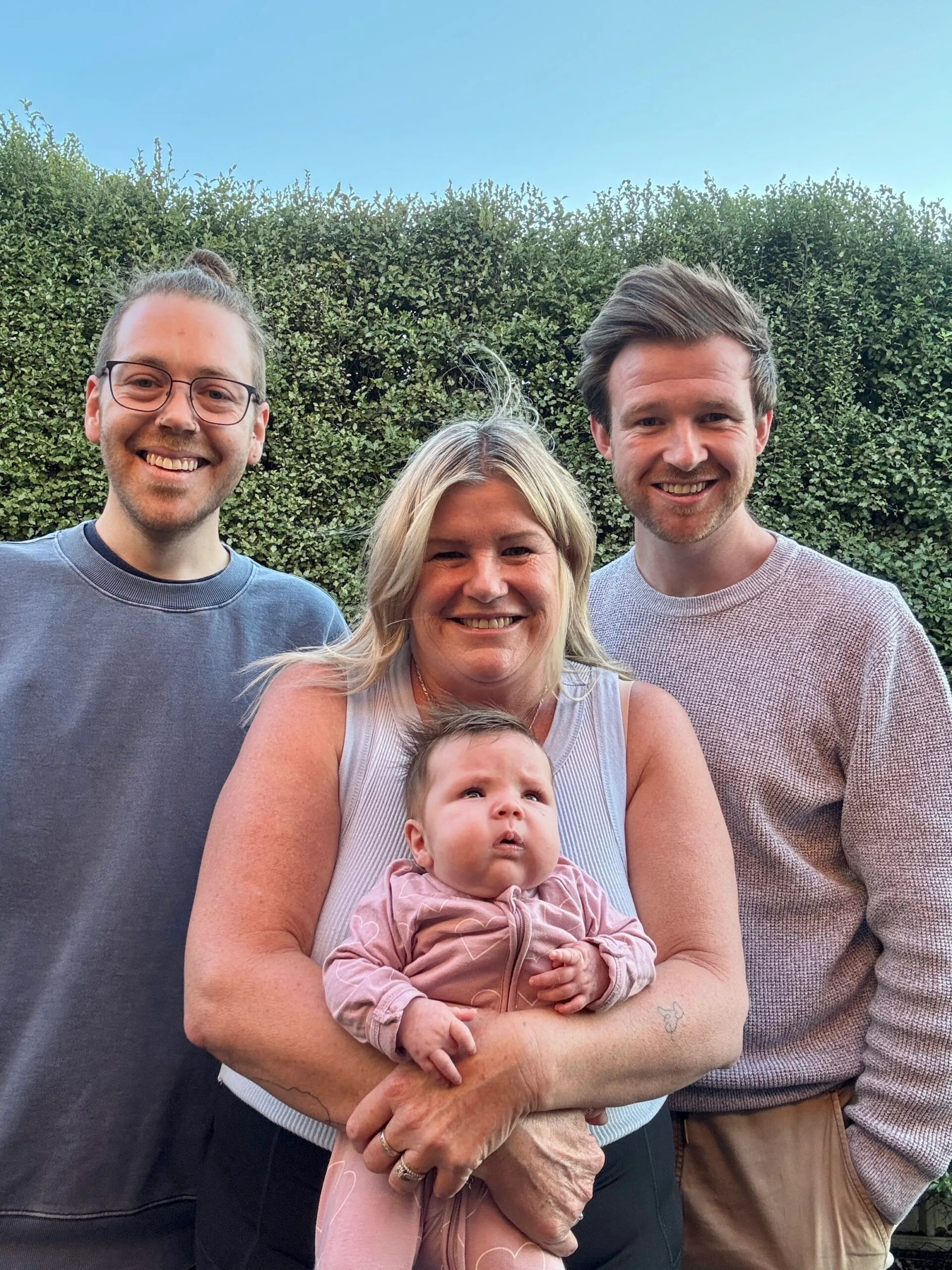 A woman stands in the middle holding a baby. She is wearing a white singlet and the baby is in a pink onesie. Her sons stand either side of her. They are both wearing jumpers