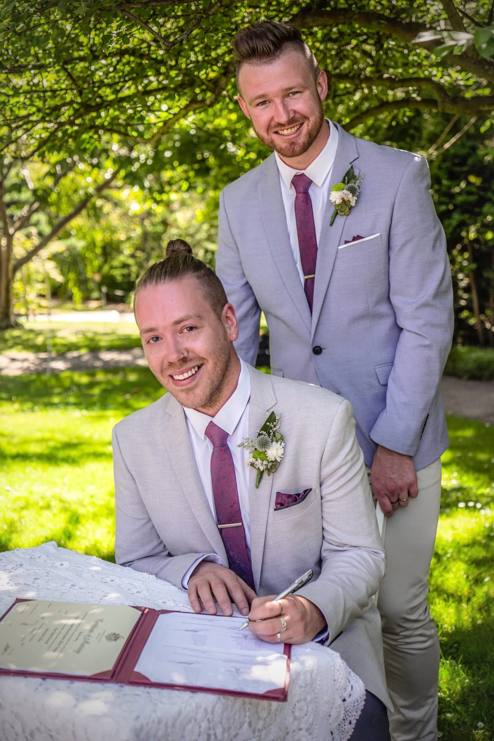 Two men on their wedding day. One is wearing a beige suit with red tie. The other is wearing a grey suit with red tie