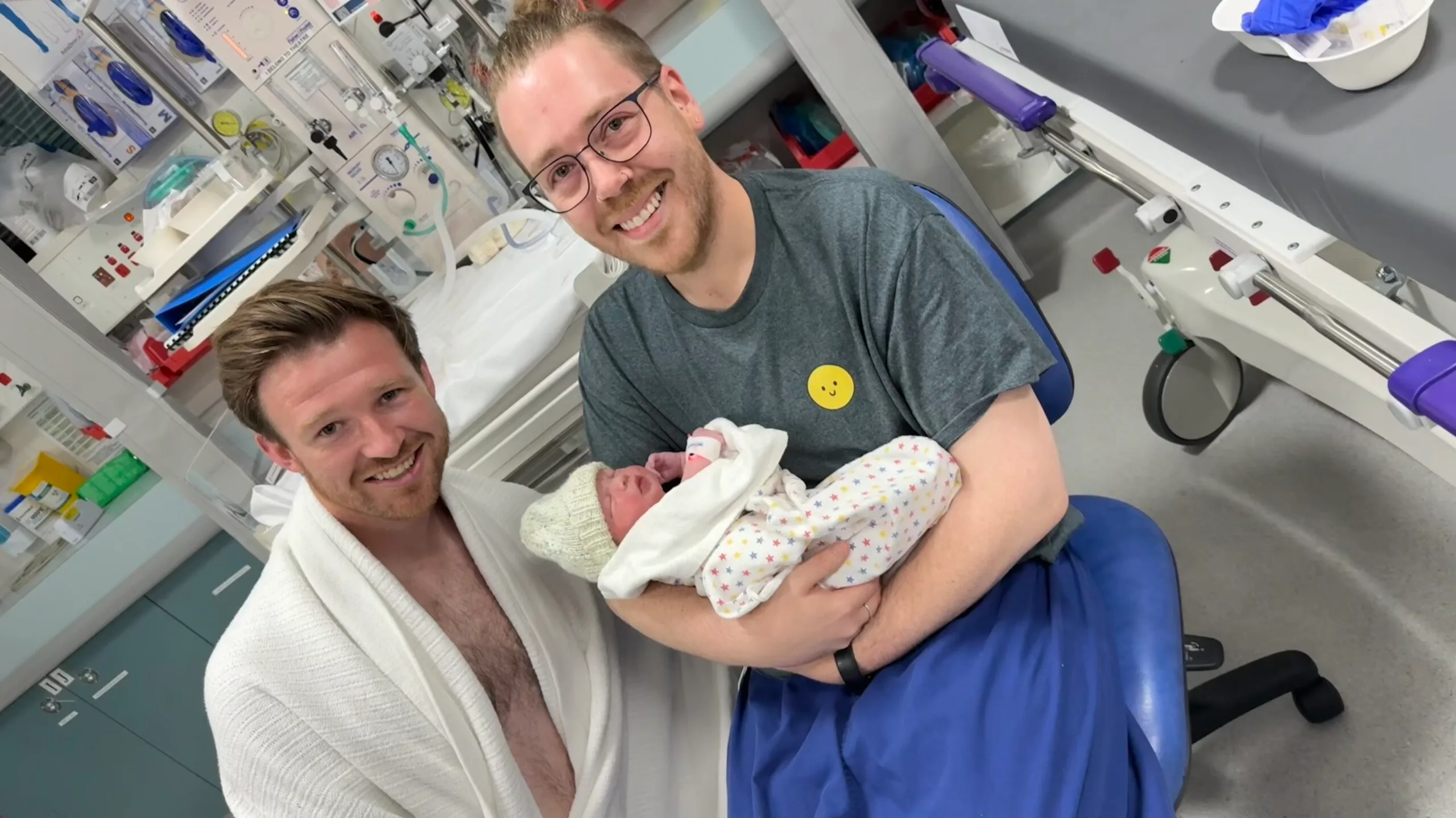 Two men hold their baby for the first time in hospital. One is wearing a white dressing gown. The other is wearing a grey shirt and blue pants
