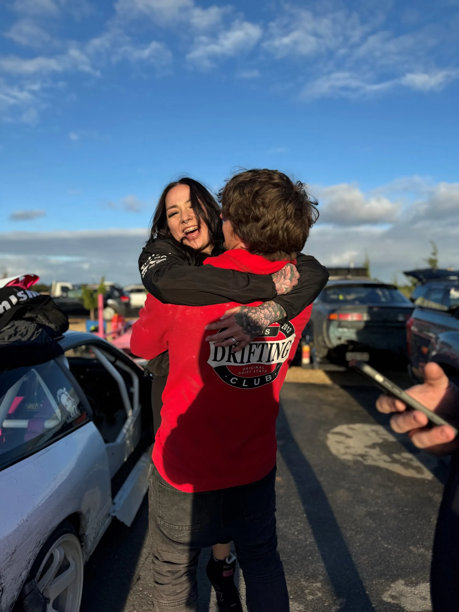 A man and woman beside a car, he is facing away from the camera picking her up in his arms she is smiling widely