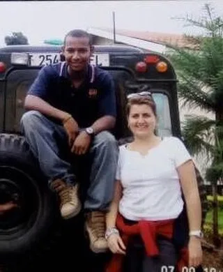 Nas sitting on a car bonnet and Donna standing next to him. Both are smiling and looking at the camera.