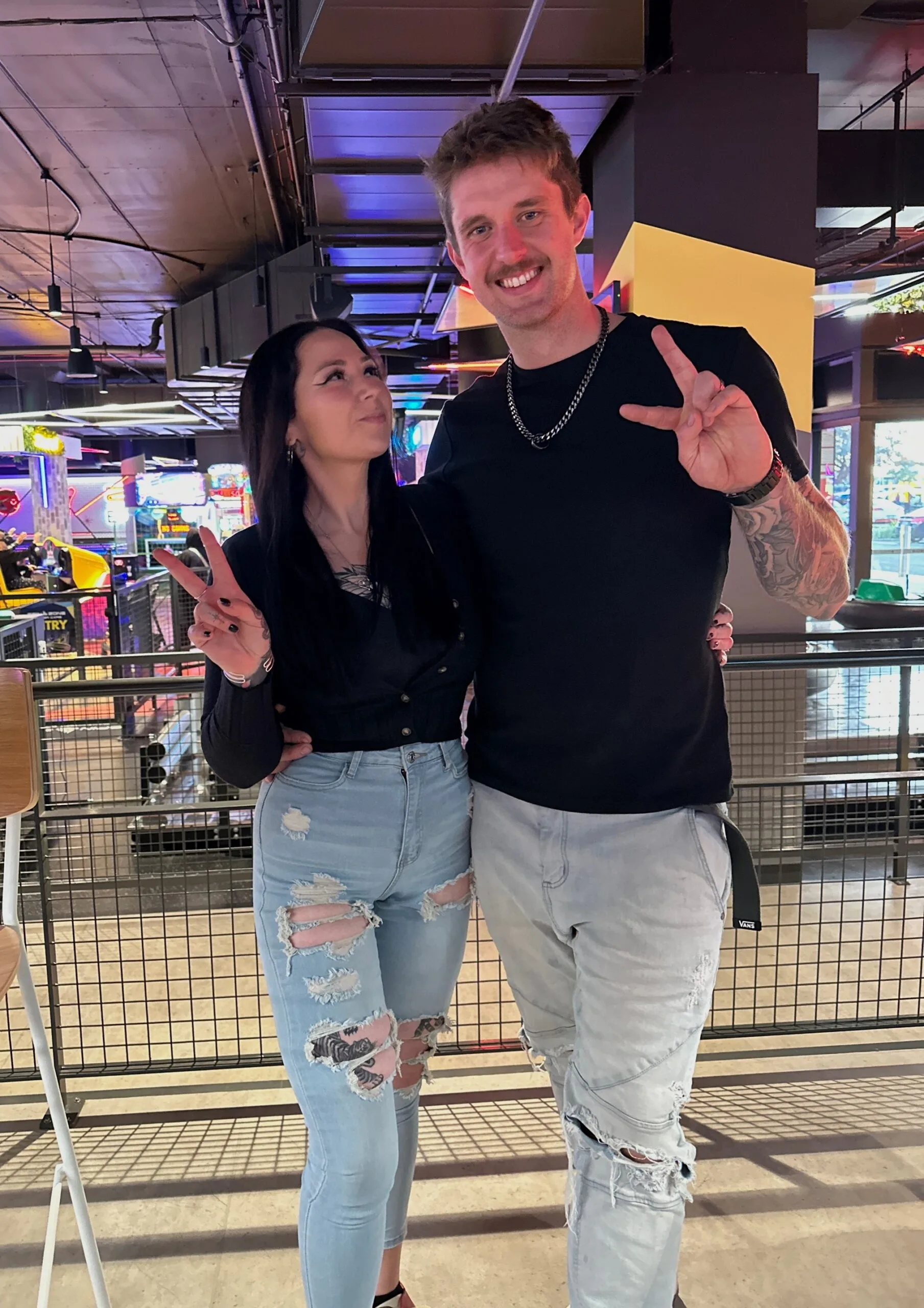A man and a woman both in jeans and tshirts at a bowling alley. she is smiling and looking at him, he is looking ta the camera and making a peace sign