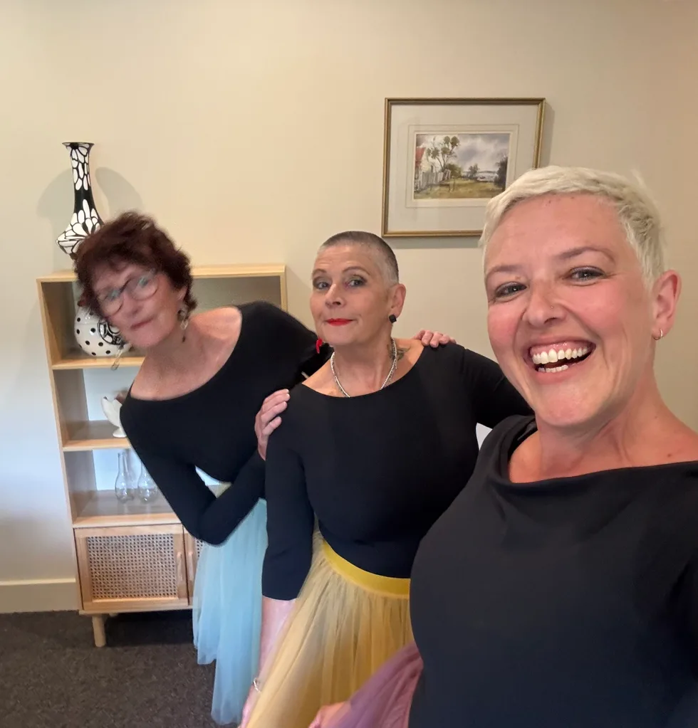 Three middle-aged women in ballerina costumes, smiling at the camera