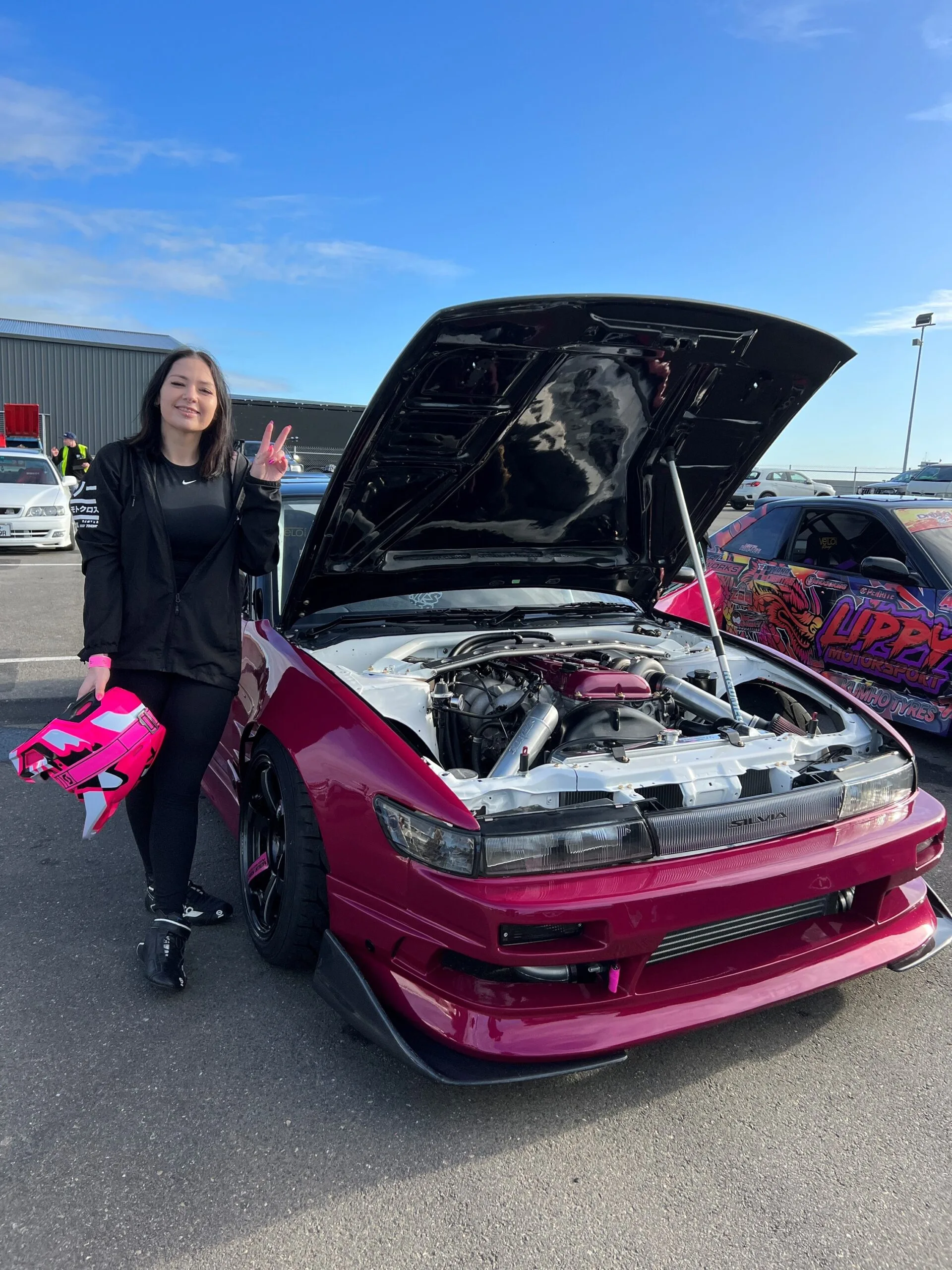 A woman wearing all black holding a pink driving helmet stands next to a maroon-pink racing car