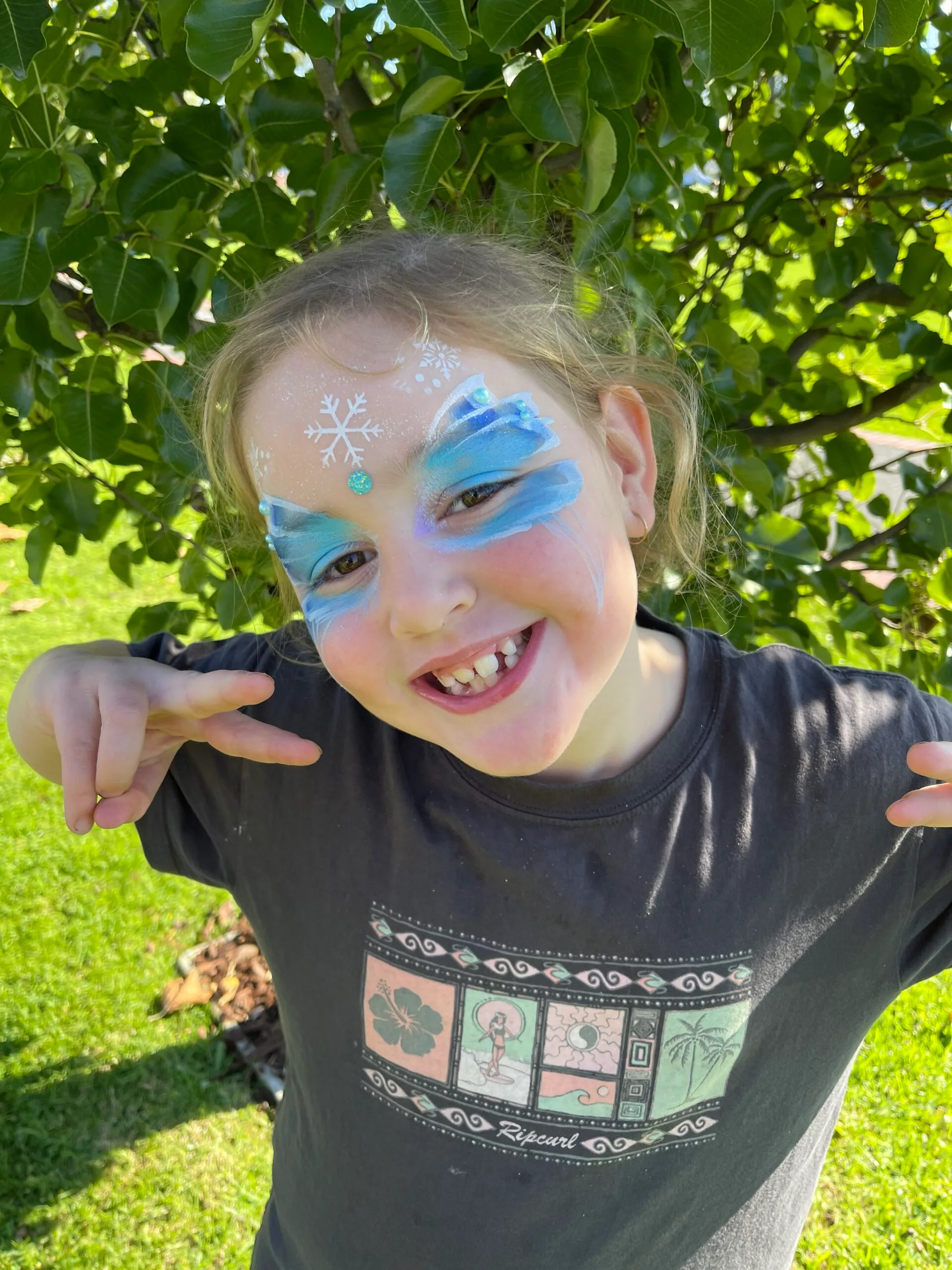 A little girl smiling in the backyard she is making two peace signs with her hands and wearing blue glittery face paint