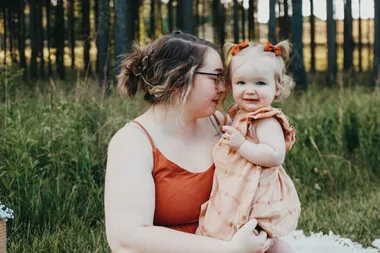 Mum in rust coloured dress holding her daughter with a peach coloured dress. They are sitting in a field on a white picnic rug