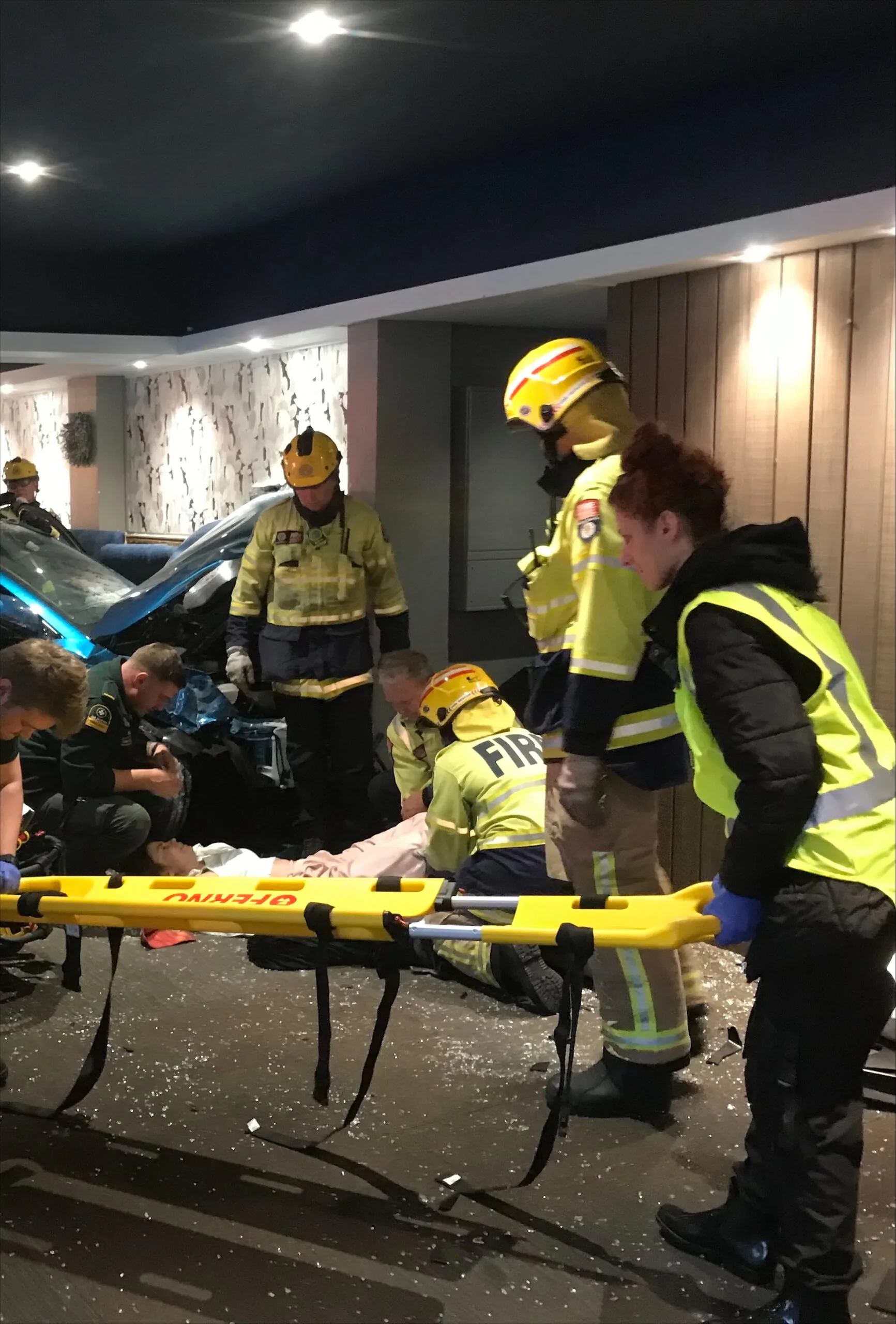 A woman lays next to a car on the floor of a restaurant, many firefighters sand around her and two are preparing a spinal board there is glass shards all over the floor
