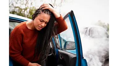 A woman steps out of a car, wincing in pain as she clutches her head.