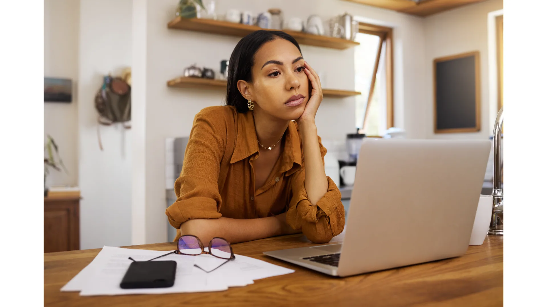 A woman sitting at a laptop stares blankly beyond the screen.