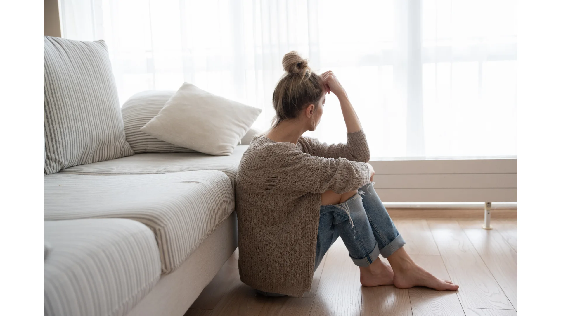 A woman appears melancholy as she stares out the window while sitting on the floor.