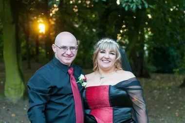 man and woman in red and black stood outside in fron tof trees.