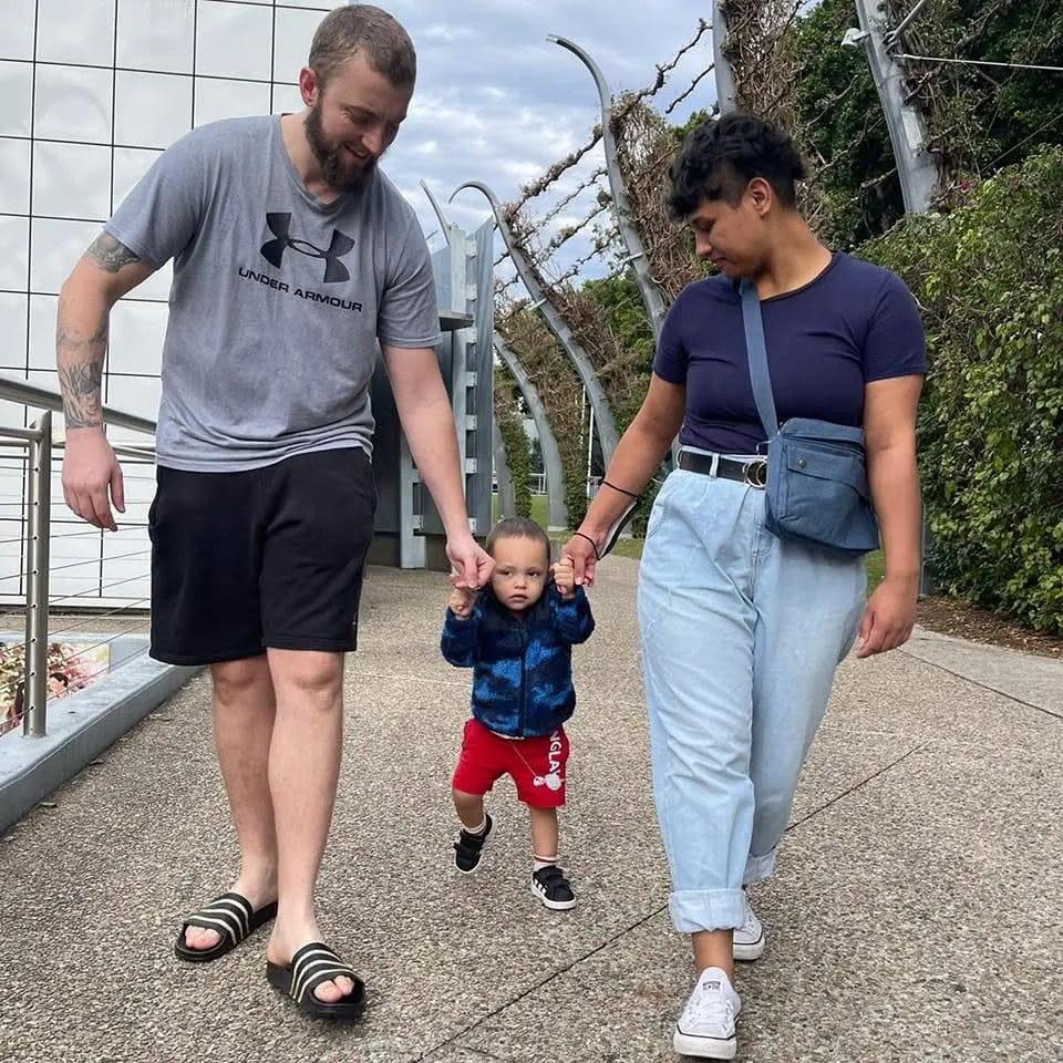 dad on left holding babies hand while walking, mum on right. 