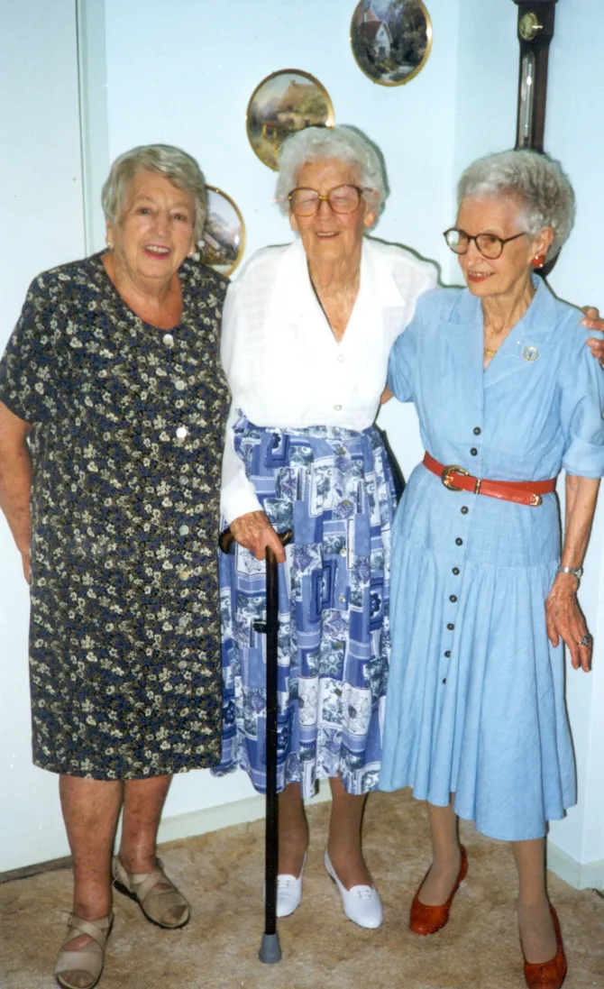 three women one wearing blue dress, one in white shirt with walking stick and one in flower dress