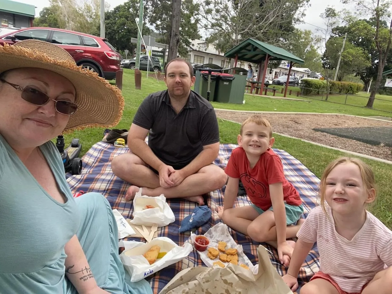 A family having fish and chips at the park. the children, a boy and girl are smiling for the camera. The mother is wearing a blue dress and the father is wearing grey. The woman has a hat and sunglasses.
