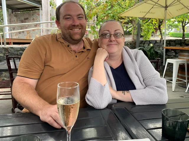 man and woman sit outside at a table drinking sparkling wine, she is wearing a grey jumper and his is wearing brown