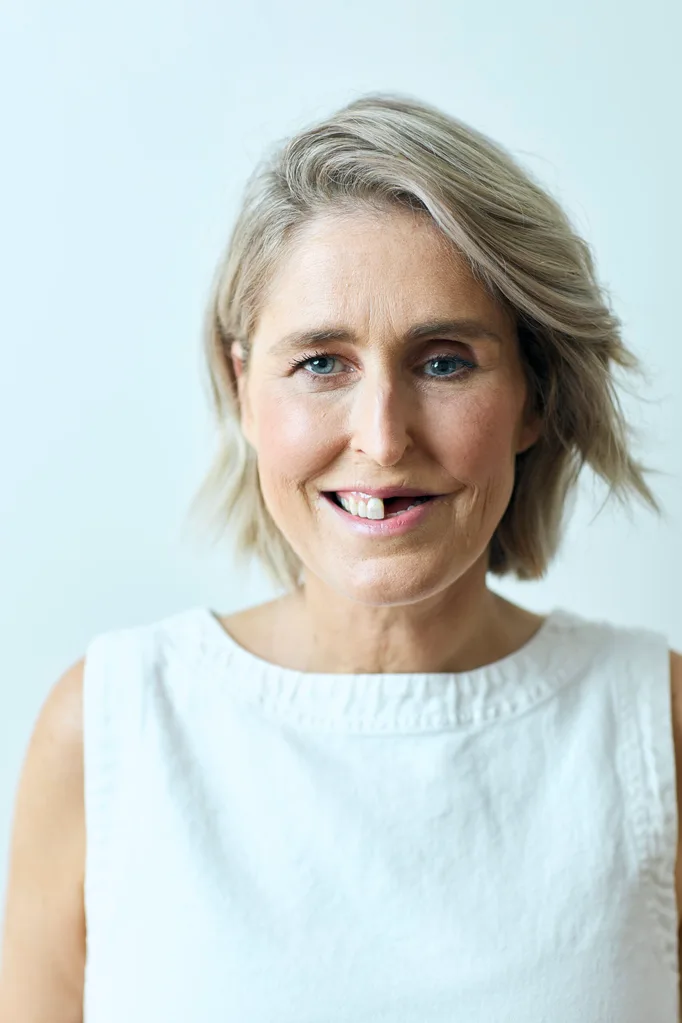 woman with blonde hair blue eyes and only half her teeth in a white top .