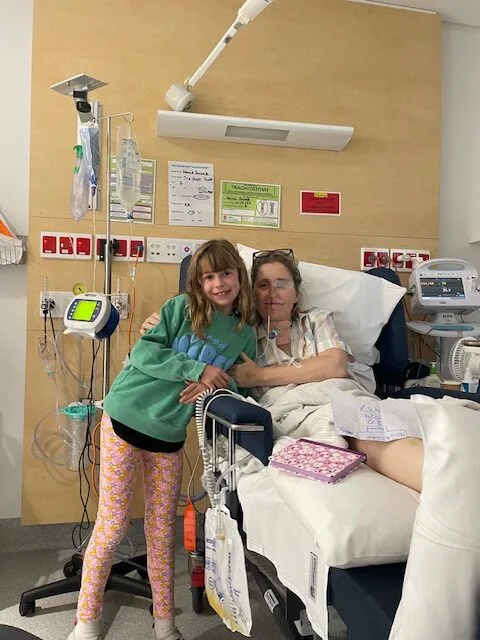 Girl in green top and black shorts and woman in hospital gown in hospital bed against white wall.