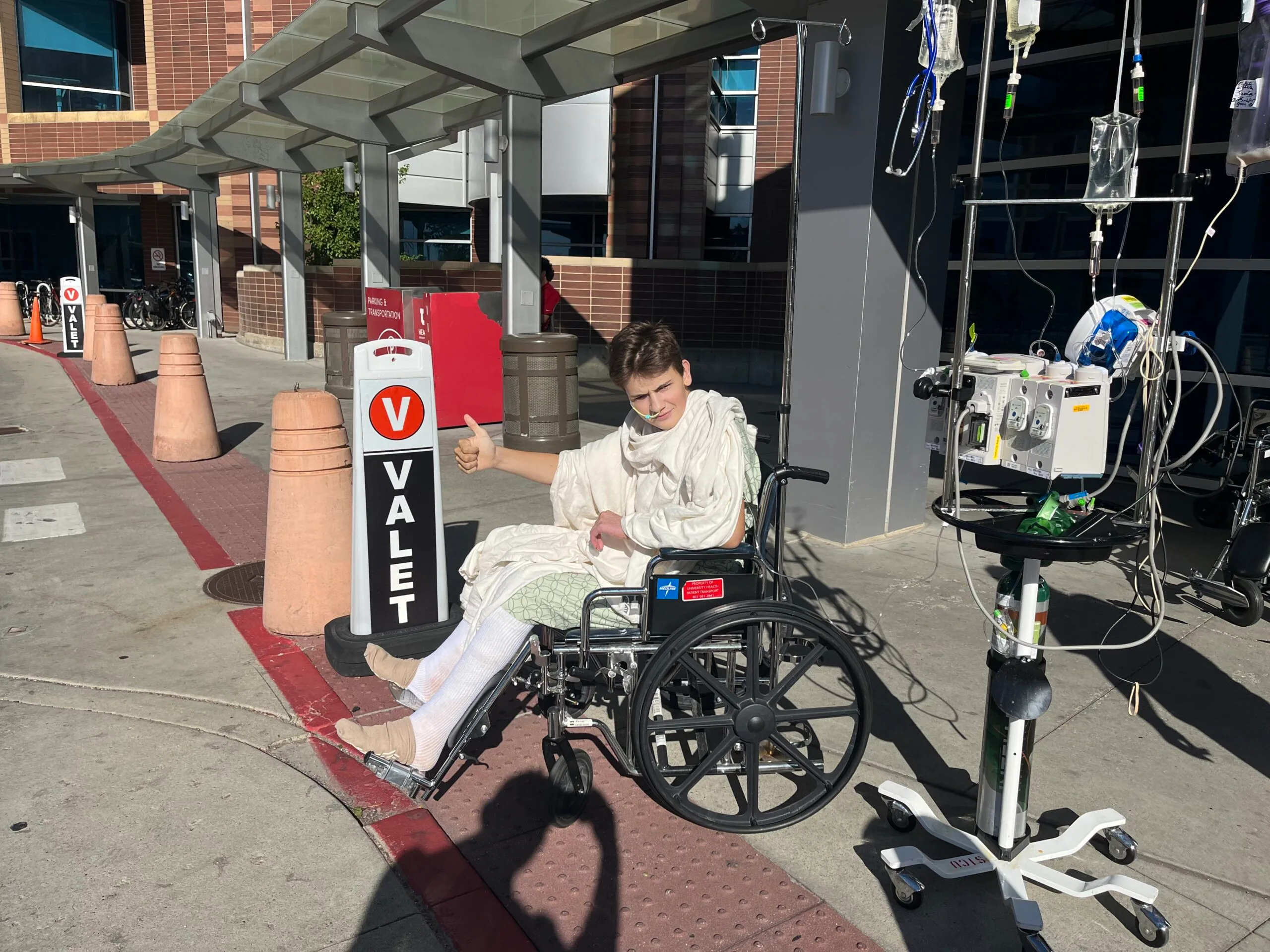 boy wrapped in bandages sitting in wheelchair outside, he is giving a thumbs up