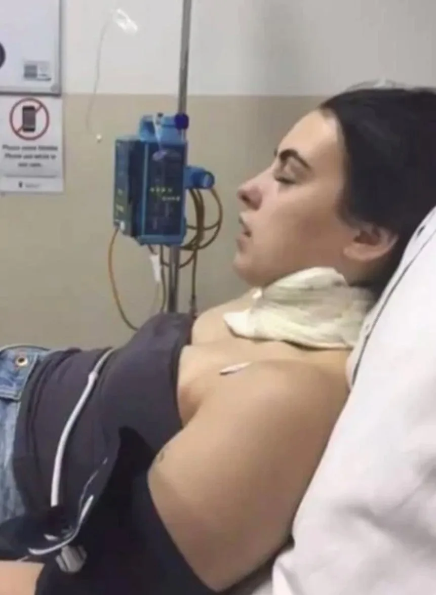 a woman sitting in a hospital bed, side profile, she has a large bandage on her neck