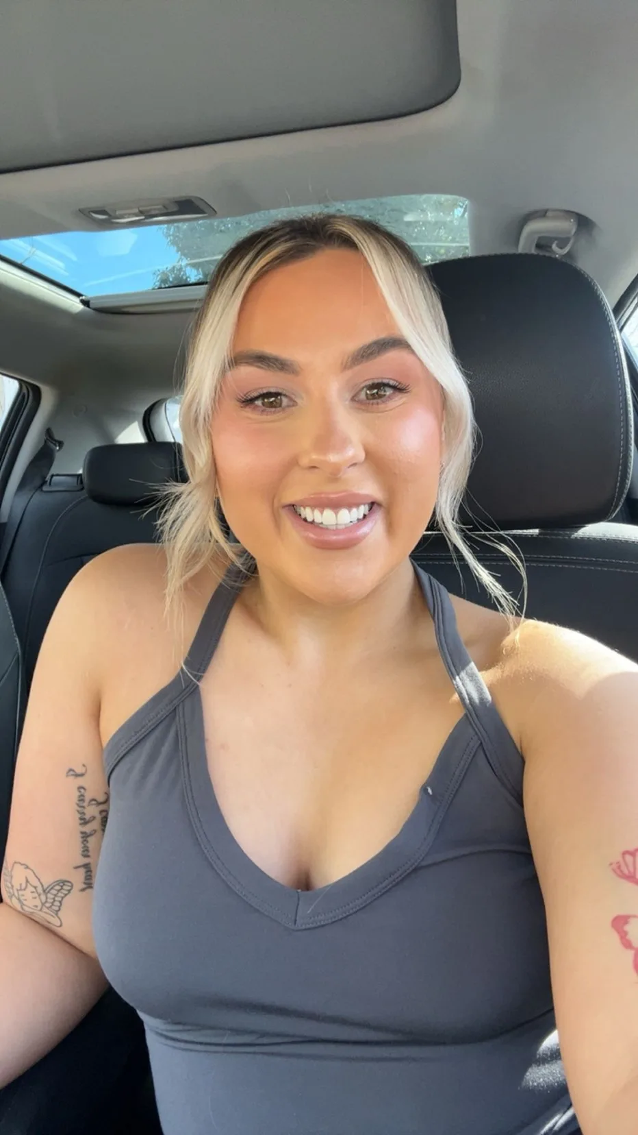 a blonde woman takes a photo of her in the car she is wearing a grey top and has brown eyes