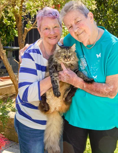 Woman on left has pink short hair and is wearing striped blue and white shirt smiling at camera holding cat. 
Woman on right has short grey hair wearing blue shirt with arms around woman next to her and holding cat.