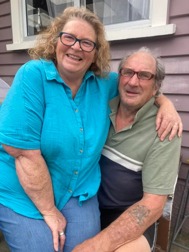 Elderly woman in blue shirt sitting on lap of elderly man in green shirt and both smiling.