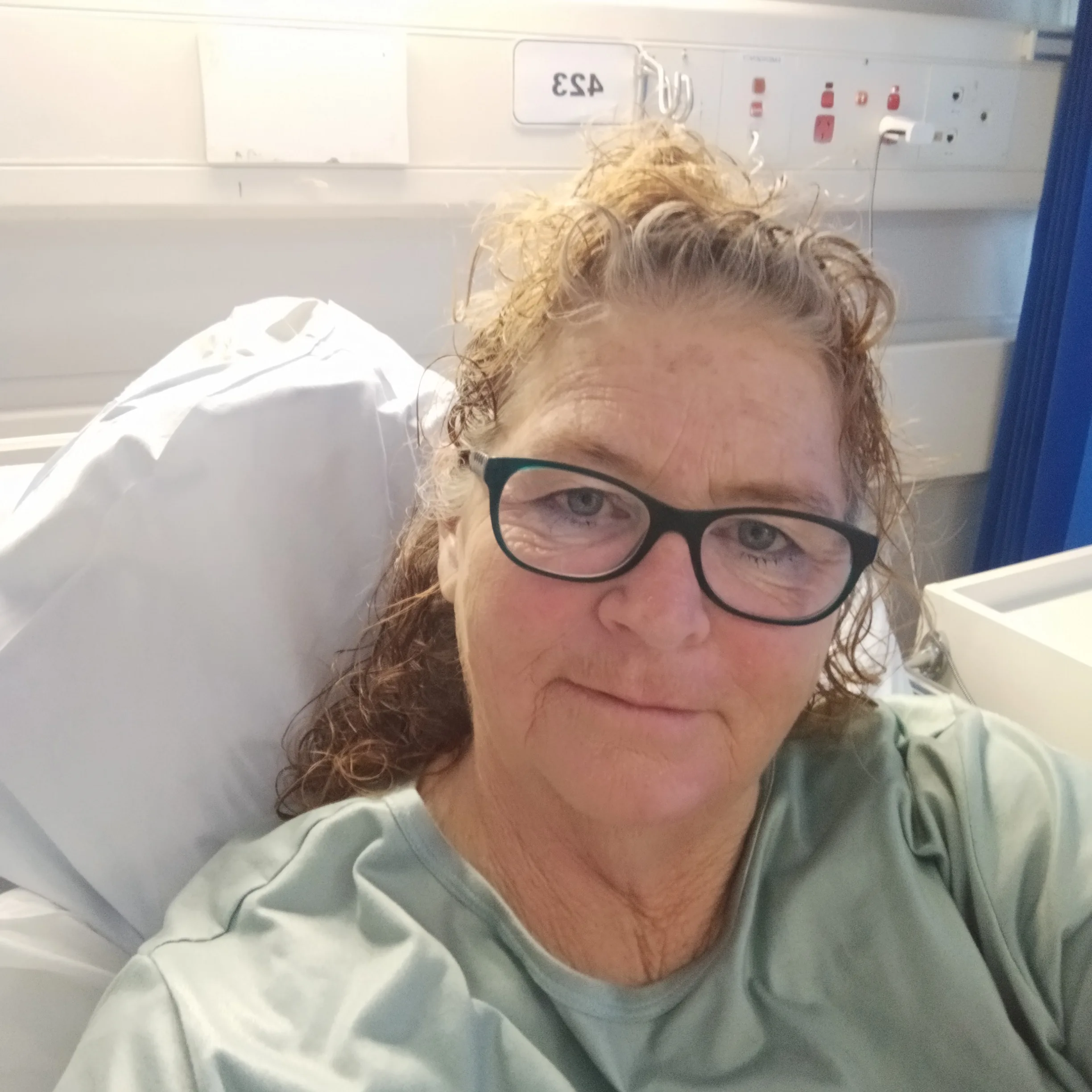 Elderly woman wearing black glasses in hospital bed looking at camera.