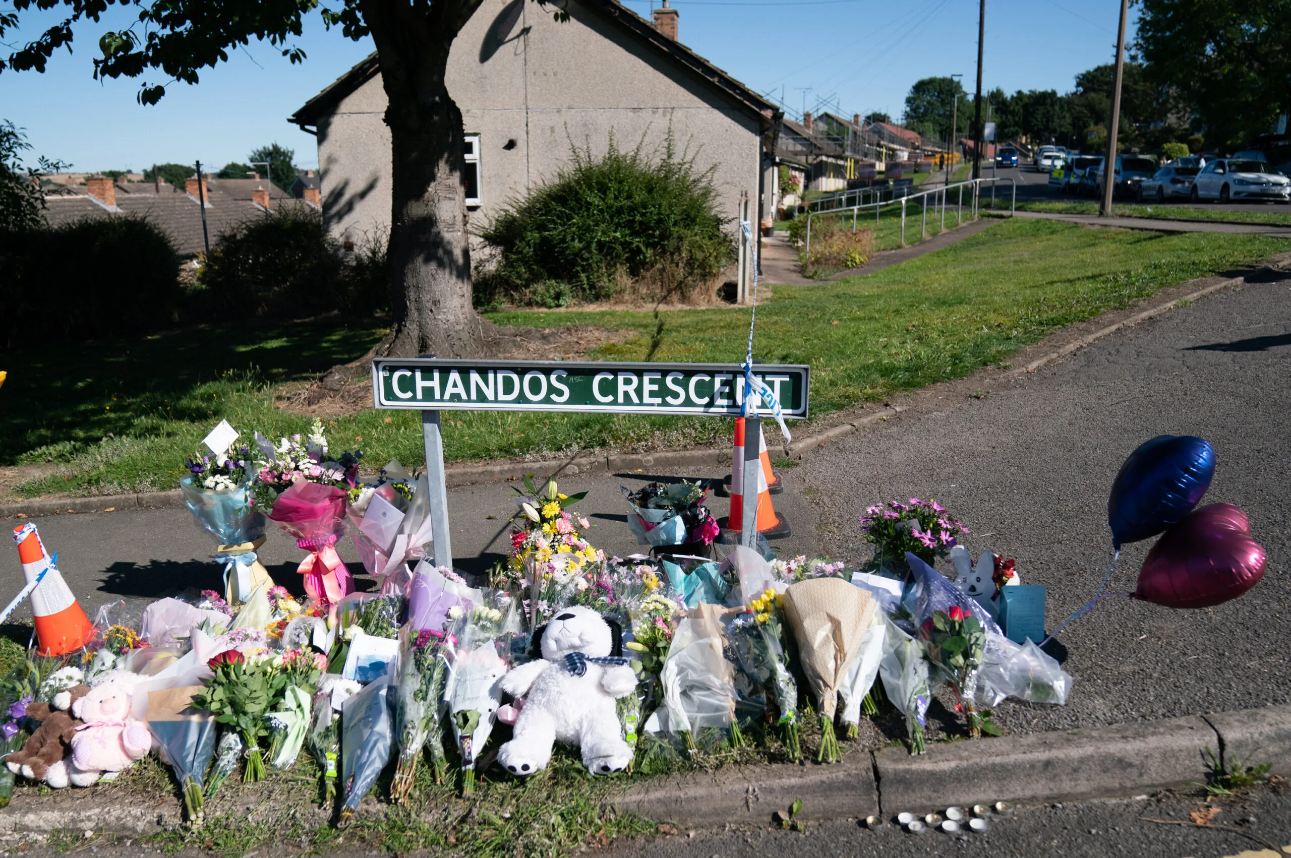 A row of flowers, soft toys and balloons in front of house.