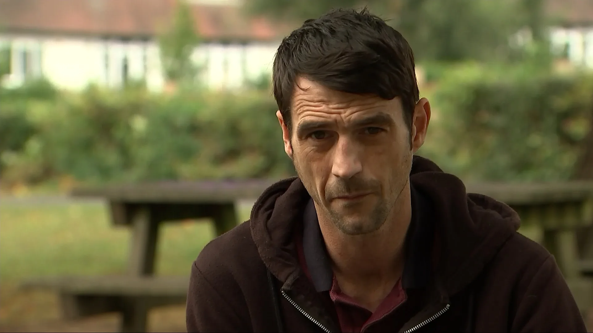 Man with black hair and dark brown jacket looking sad on camera.