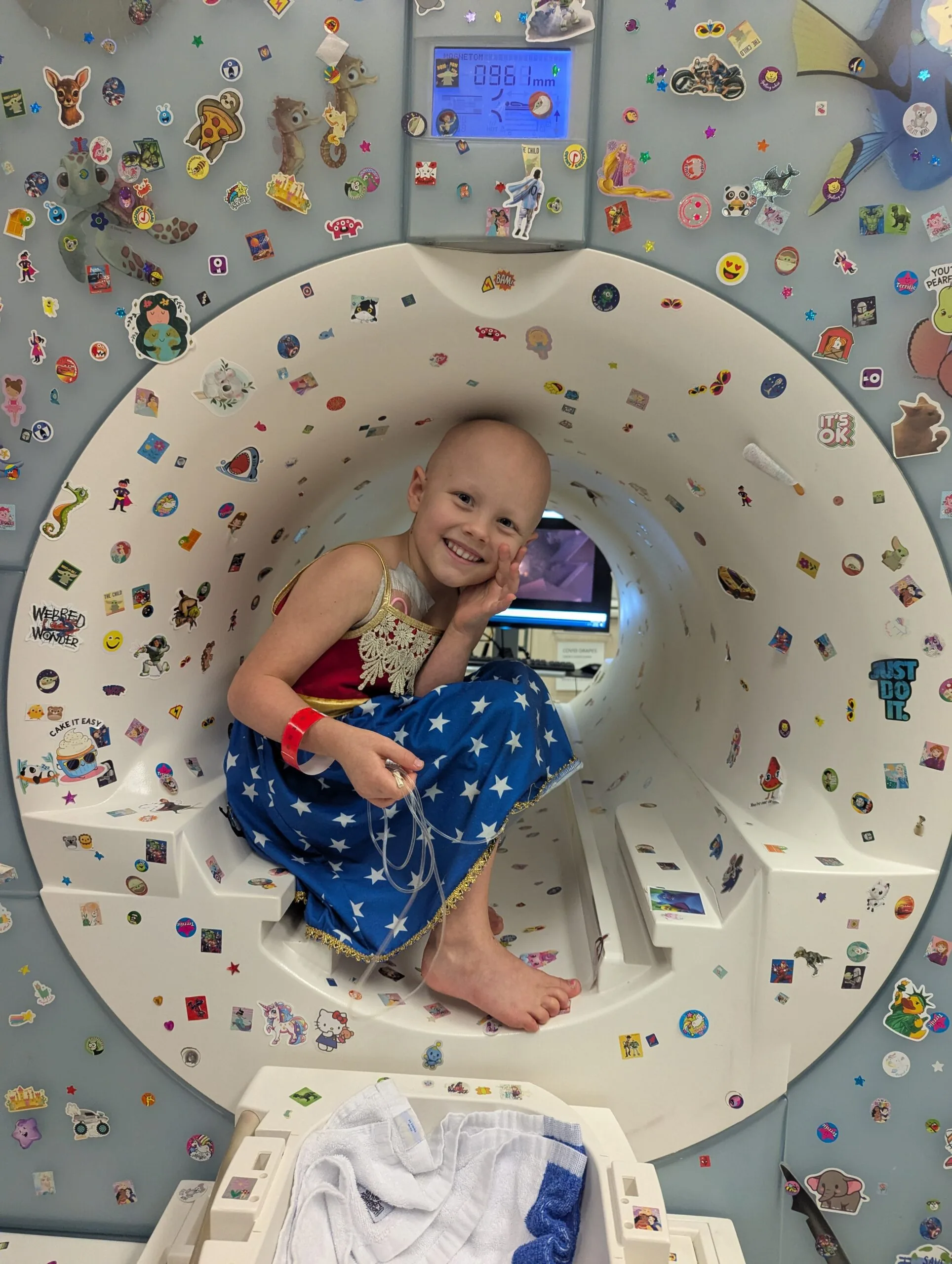 a young girl with no hair dressed as wonder woman in an MRI machine covered in stickers