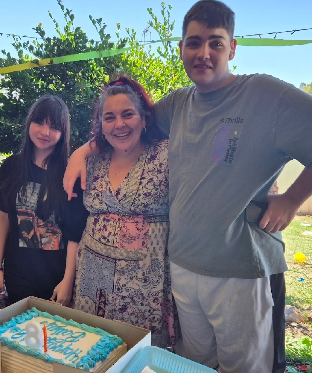 two young adult children with mother in the middle in front of blue iced cake