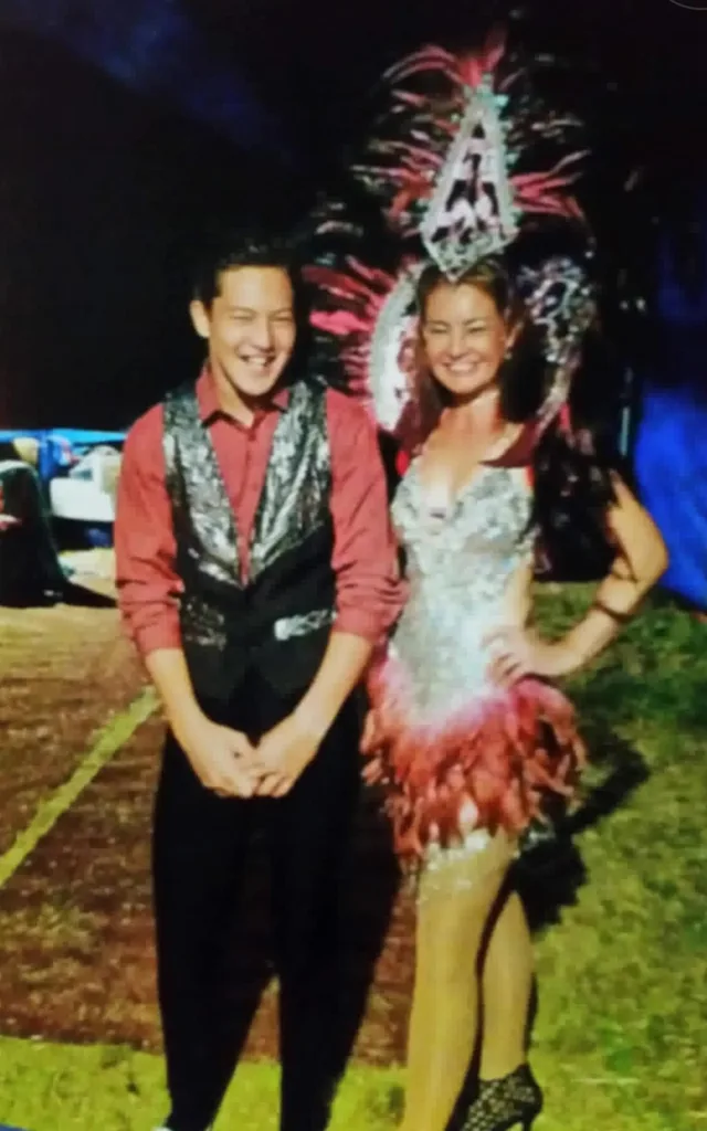 Woman in sparkly show girl dress with pink feathers and headress and boy with pink shirt and black suit standing