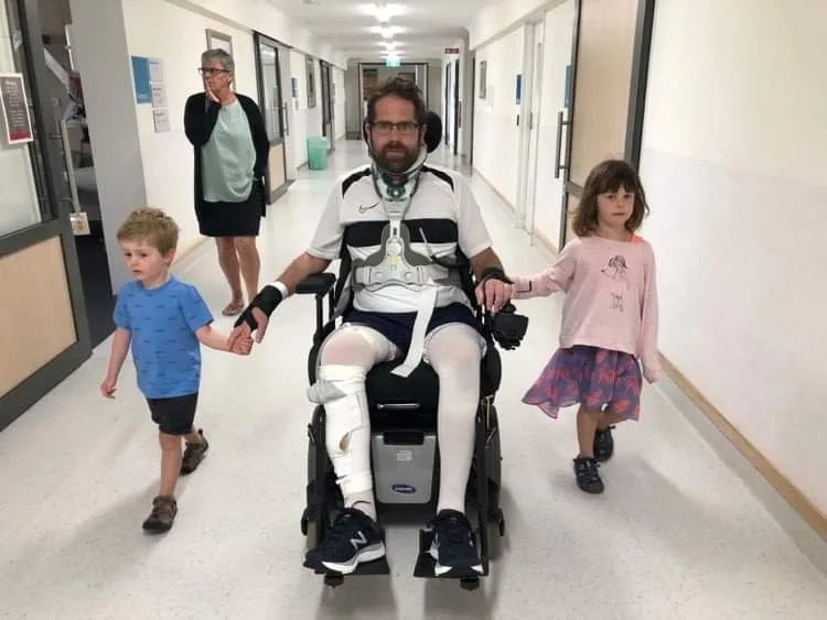 Dad in wheelchair, on left young boy holds his hand he's wearing blue and little girl on right is wearing pink