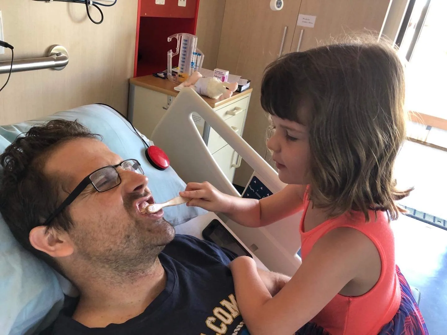 a young girl in orange sits with dad in hospital bed brushing his teeth