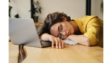 A woman sleeps at her desk, slumped over her laptop.