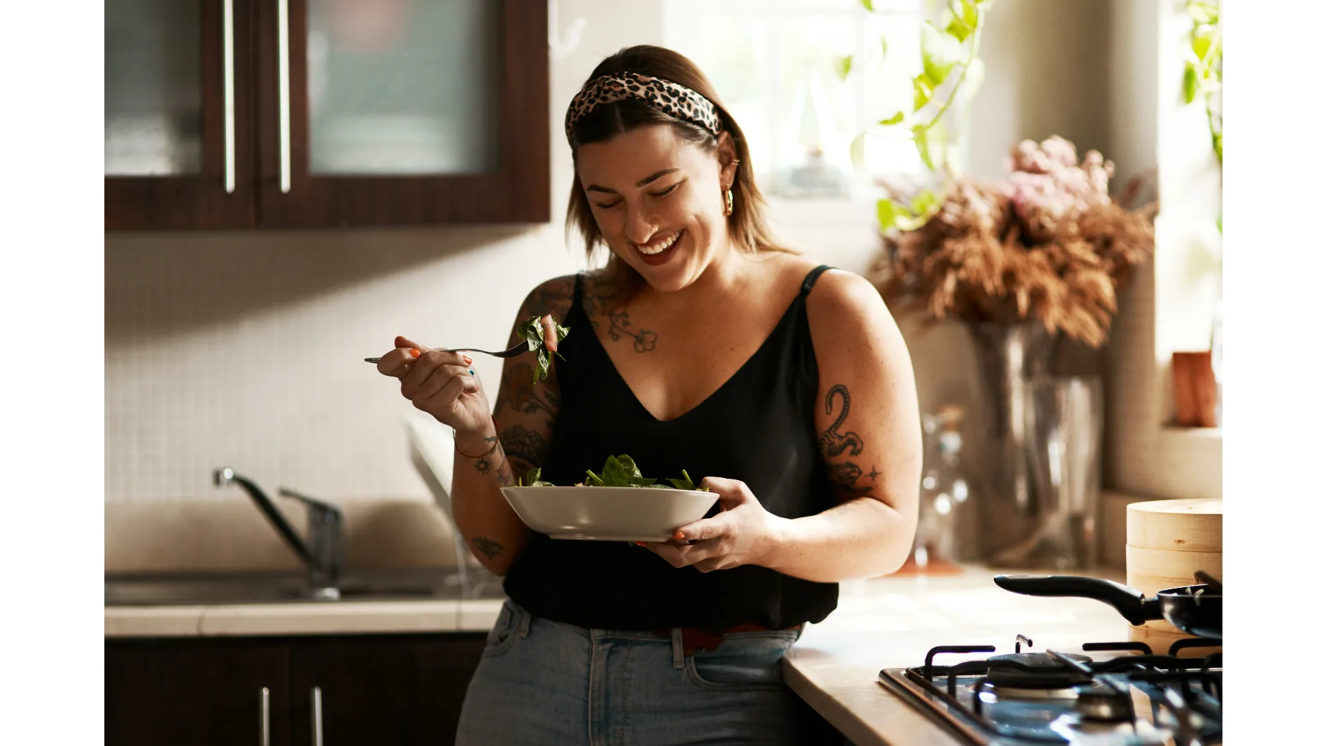 A young woman stands in a home kitchen, smiling as she eats a nutritious bowl of food.