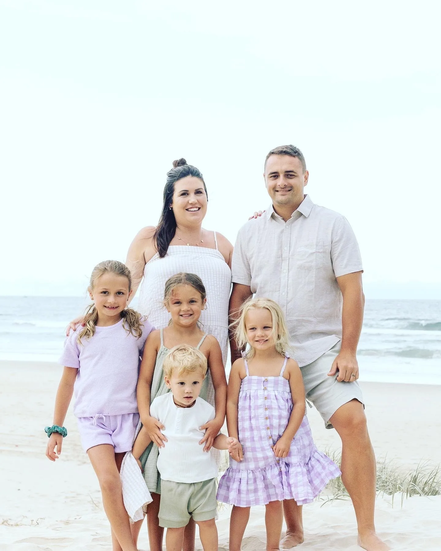 Woman with dark hair wearing white dress standing next to man wearing white shirt with short hair. 

Three young girls standing in front of man and woman on the beach holding on to toddler boy with blonde hair. 

All smiling at camera. 