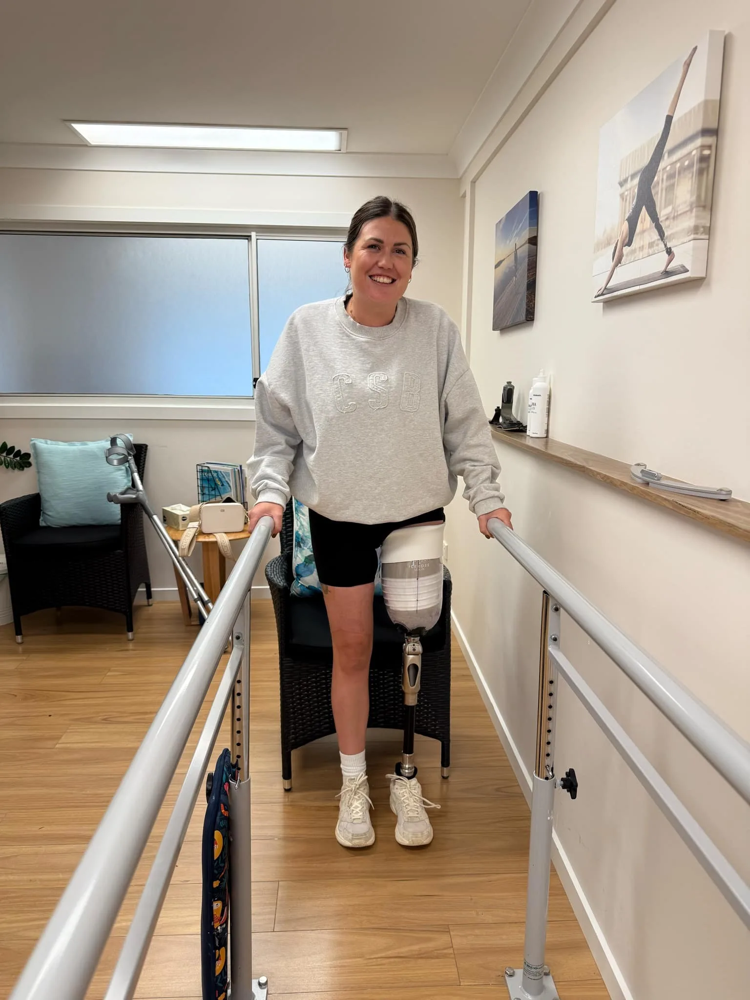 Woman wearing grey jumper and black shorts trying on prosthetic leg on left side while holding on to railings. Smiling at camera. 