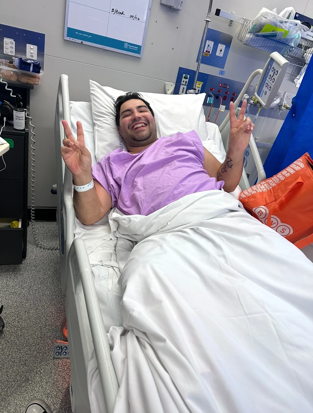 man in purple scrubs in hospital bed throwing peace signs