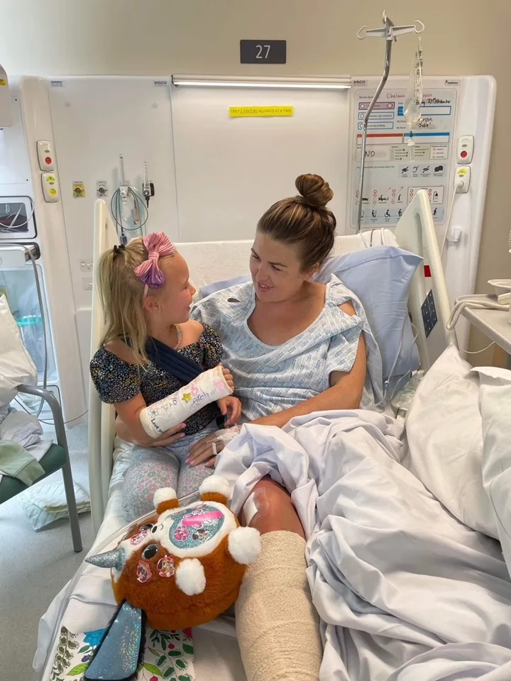 Little girl with blonde hair and fractured arm sitting next to woman with bandaged leg on hospital bed. Both smiling at each other.