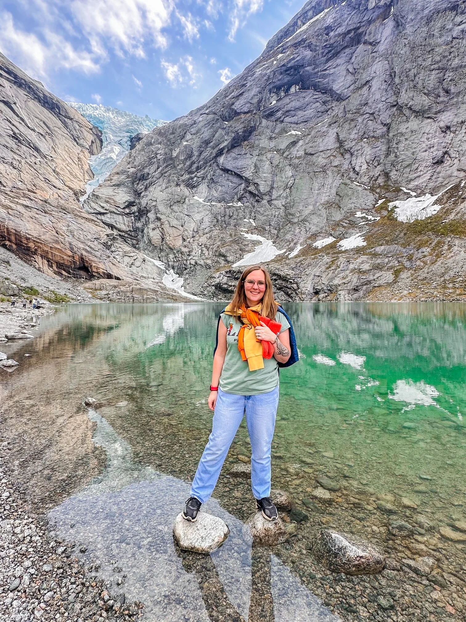Woman wearing green shirt, blue jeans and glasses, standing on top of rocks in a lake smiling at camera. 