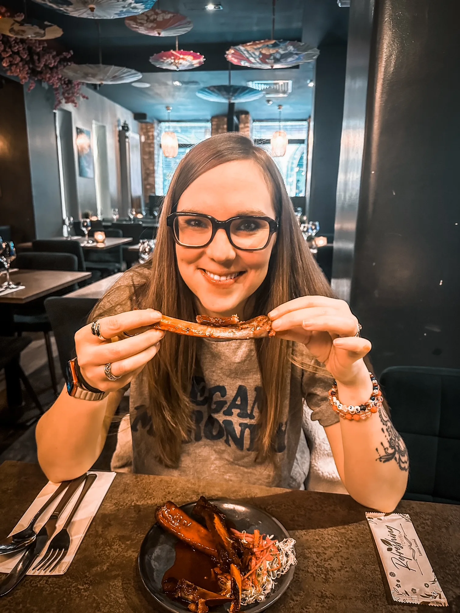 Woman with long brown hair wearing black glasses holding up singular beef rib and smiling at camera at restaurant. 