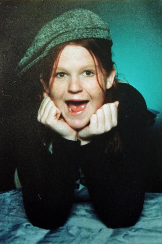 Young girl with red hair, wearing grey beret, resting head on hands and smiling at camera.