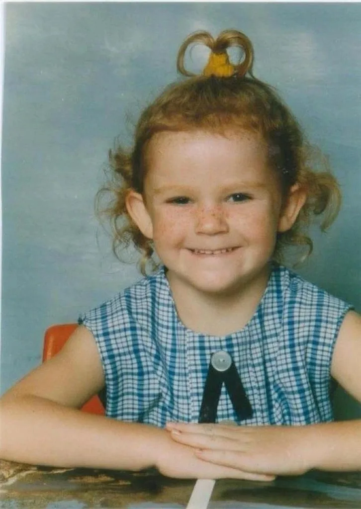 Baby girl with freckles across her face and red hair, wearing blue and white dress, smiling at camera.