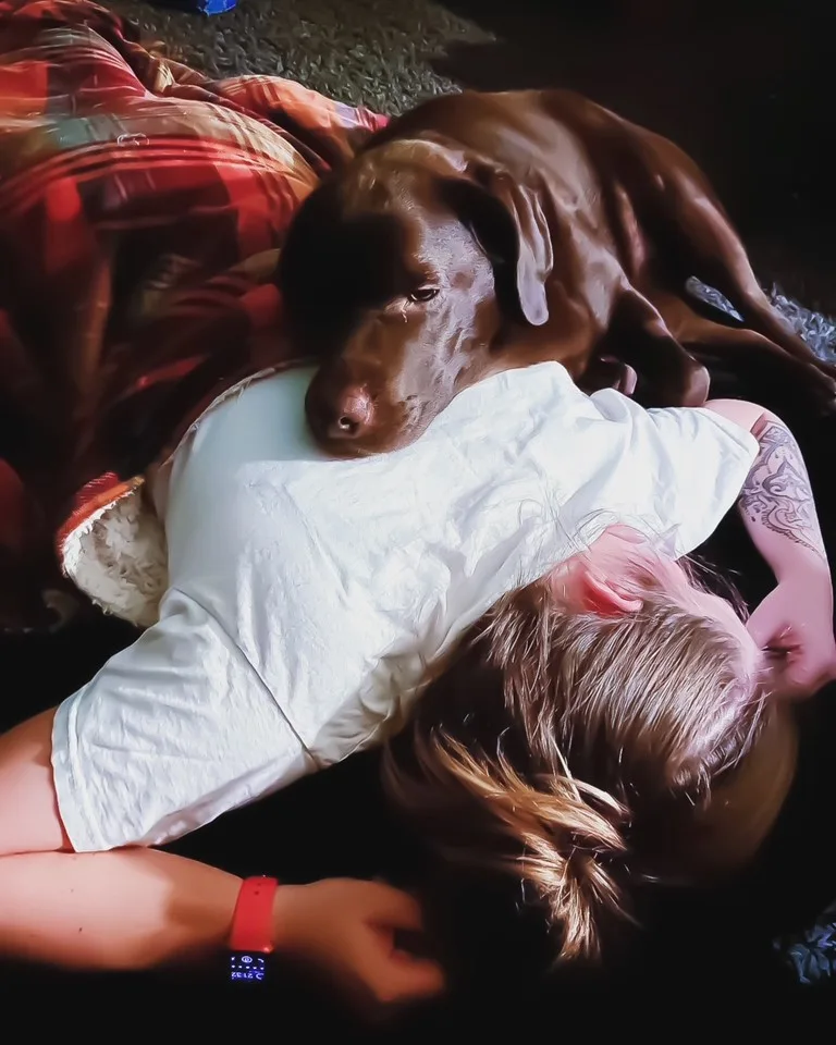 Woman lying face down on floor covered by blanket as brown dog rests itself on her back.