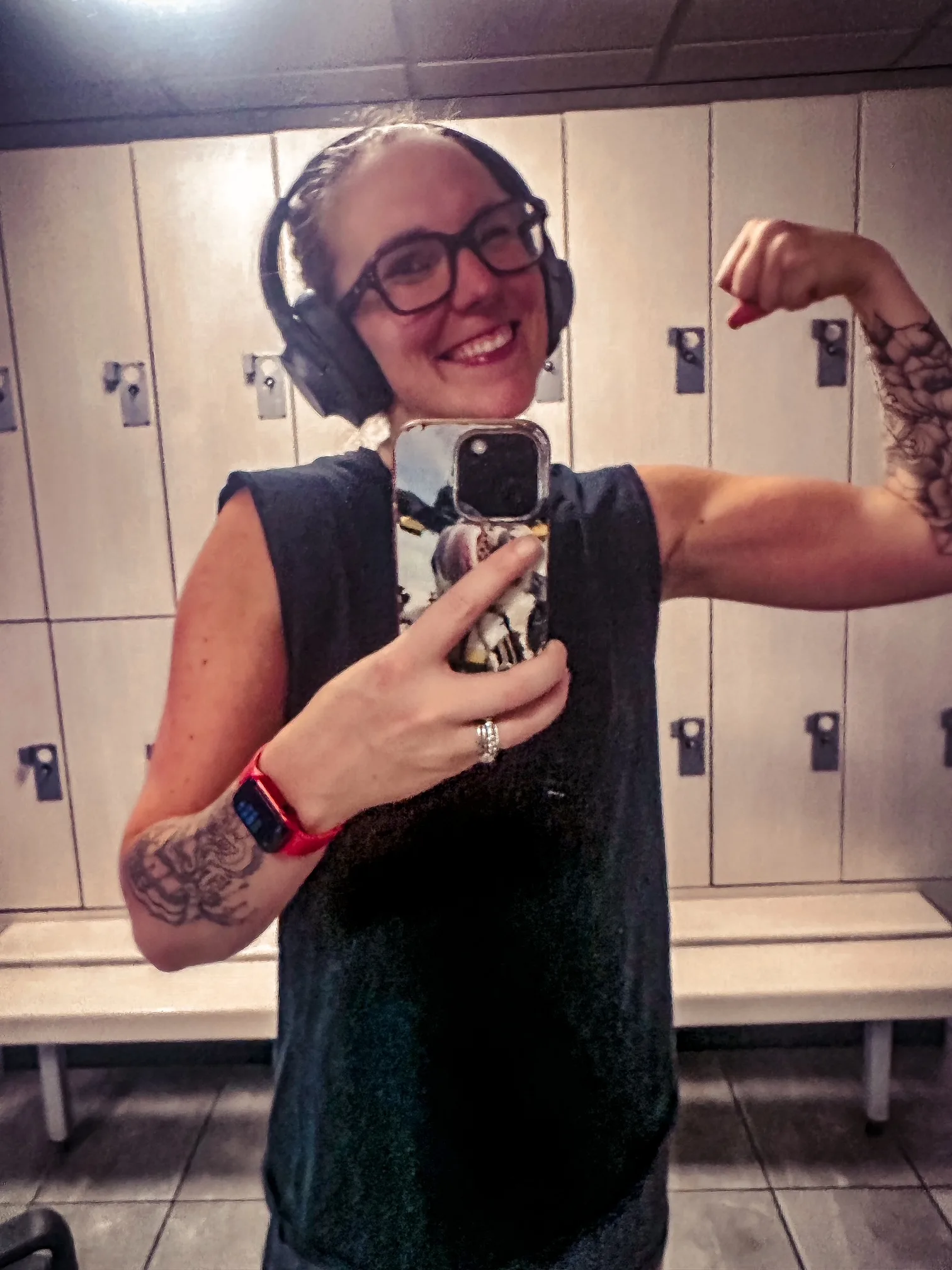 Woman wearing black tank top and black headphones, flexing biceps in mirror and smiling while taking mirror selfie. 