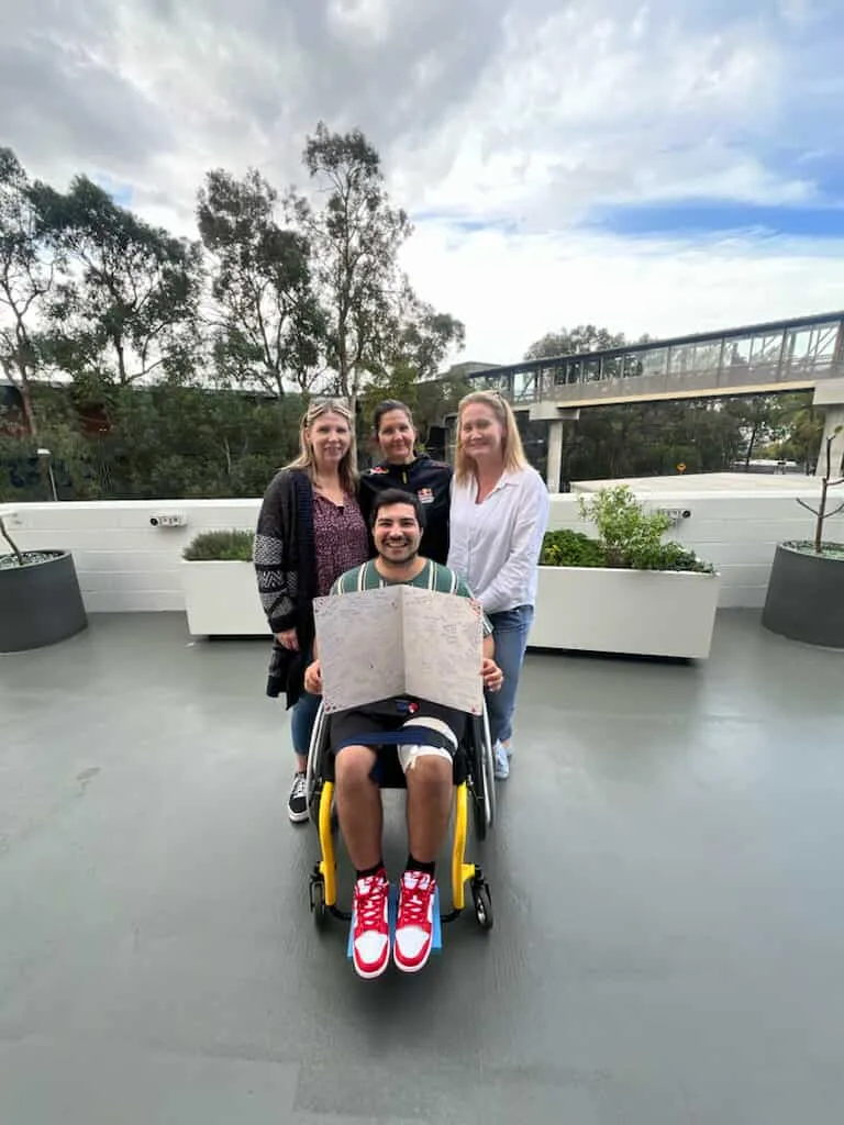 man in wheelcahir with three females friends he is holding a big card