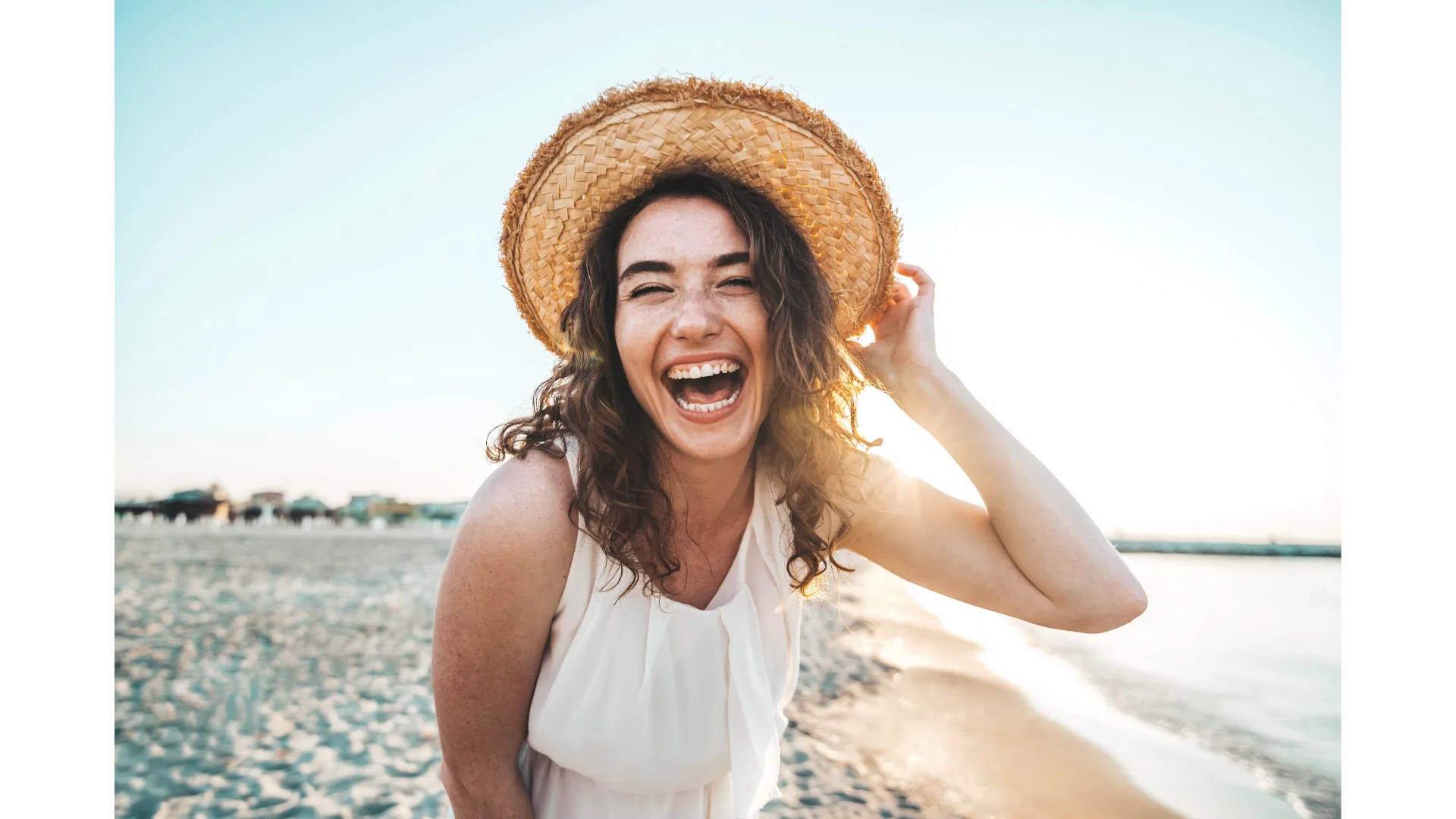 A young woman holds onto her hat while laughing on a beach.