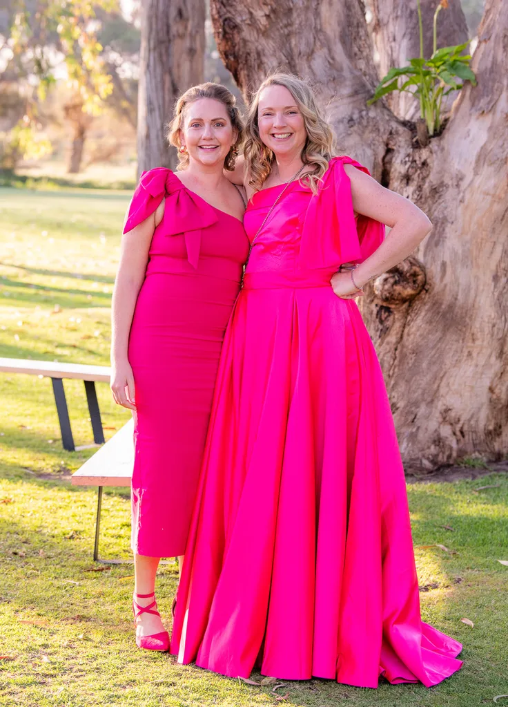 two women in pink dresses stood in front of tree and bench on grass